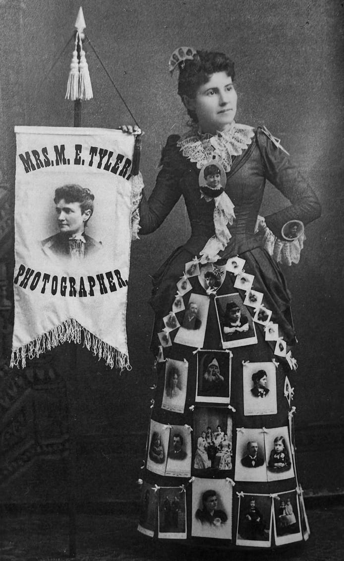 1892 marketing dress for Mae Tyler, photographer. Mrs. Tyler's photo is on the banner. Dress is worn by her assistant, Cora Baldwin.
