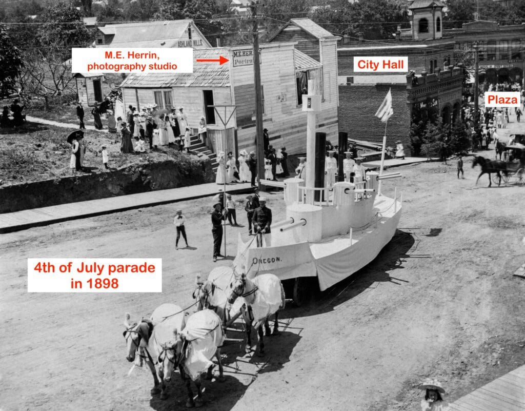 Photo of USS Oregon float in 1898 4th of July parade, during the Spanish-American war. Note sign for M.E. Herrin Portrait photographer on building.